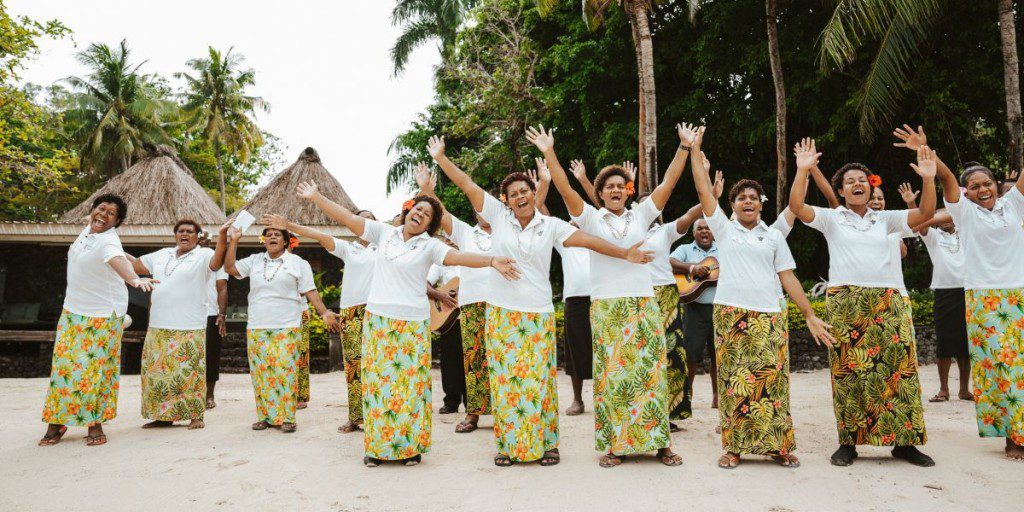 A group of women dressed in vibrant, floral-patterned skirts and white shirts, joyfully performing a dance on a sandy beach with tropical huts and lush greenery in the background.