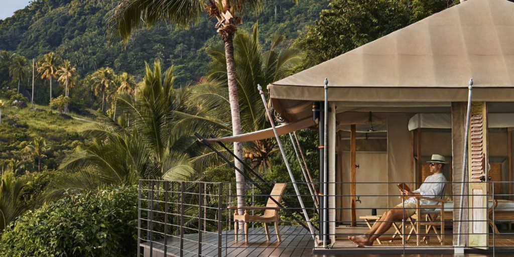 A man sitting relaxed on the deck of a luxurious tented accommodation, surrounded by tropical palm trees and lush greenery, with a mountainous landscape in the background.