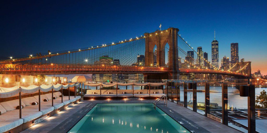 Scenic view of an illuminated rooftop pool and seating area overlooking the Brooklyn Bridge and the New York City skyline at dusk