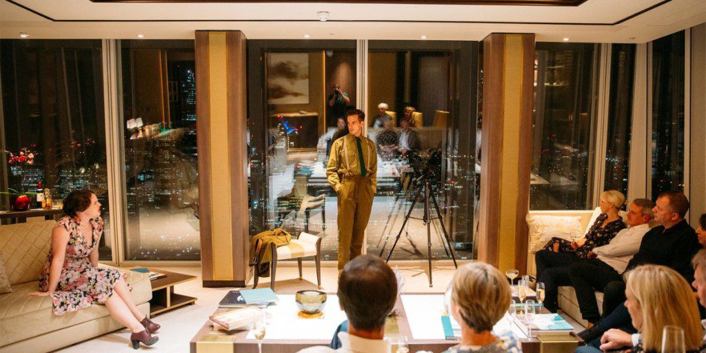 A man giving a presentation to a small group seated in a modern, upscale room with floor-to-ceiling windows showcasing a nighttime city skyline